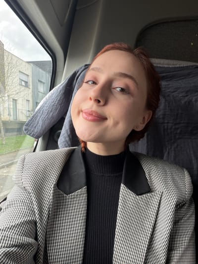 A female individual is depicted in a close-up, chest-up shot from inside a vehicle, in what is identified as Eschau, France. She has reddish-brown hair and light skin, with dimples visible on her cheeks, and a slight smile directed at the camera. She is wearing a black ribbed high-neck top beneath a blazer featuring a black and white small-checkered pattern with distinct black, angular lapels. A small silver stud earring is visible in her left earlobe. The vehicle's interior includes a light gray quilted fabric draped to the subject's left and a dark gray textured material behind her head. Through the vehicle's window, an overcast sky is visible above an exterior scene featuring a light-colored multi-story building with a metal railing, a bare-branched tree, and green ground cover.