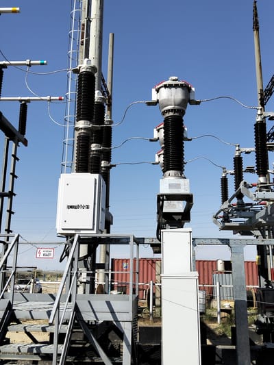 This image shows an outdoor electrical substation on a clear, sunny day, likely in the afternoon given the bright sunlight and distinct shadows. The scene is dominated by tall, grey metal structures supporting a complex network of electrical equipment. Several large, black, ribbed insulators are prominent, along with silver-colored electrical transformers and various wires and conductors.
In the foreground, a grey metal staircase leads up to a platform. A white electrical control box is mounted on a pole to the left of the staircase, and it bears text in Cyrillic script. To the left of the staircase, a red warning sign is attached to a fence. The sign features a lightning bolt symbol and Cyrillic text. In the background, a chain-link fence separates the substation from a dusty area with some sparse vegetation. Beyond the fence, there are shipping containers, one red and corrugated, and another grey, ribbed structure. A car is partially visible behind the fence.
The visible text on the white box reads "ПРИВОД В-Т-2-ПО". The red warning sign displays "НЕ ВЛЕЗАЙ УБЬЕТ!", which translates to "DO NOT CLIMB, IT WILL KILL!". The overall impression is one of industrial infrastructure in an open, arid environment. There are no people visible in the image. The clear blue sky suggests good weather conditions. The location is identified as Батыр ауылдық округі, Kazakhstan.