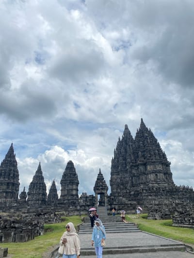 The image shows the Prambanan temple complex located in the village of Prambanan, in the Kecamatan Prambanan district, in the Special Region of Yogyakarta, Indonesia. It features a large grey stone temple with several spires, surrounded by smaller stone temples. There are several people walking up a set of stone steps towards the temple. A woman in a light blue shirt and a woman in a hijab are in the foreground, walking up the stairs. The sky is overcast, with white clouds covering the majority of the sky. There are green plants growing in the area around the temples. The steps are gray stone. The scene is calm, with a clear view of the temple complex. The temple has a lot of intricate detail in its architecture.