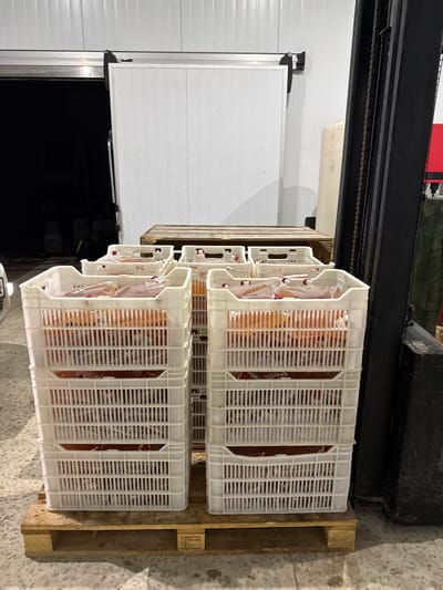 The image shows several white plastic crates stacked on a wooden pallet, containing items wrapped in plastic. The crates are filled with what appear to be food products, possibly liquids, in translucent bags with a reddish hue. The scene takes place indoors, likely in a warehouse or cold storage facility, with a large white sliding door visible in the background. A forklift is partially visible on the right side of the image, suggesting the crates are being moved or stored. The floor is concrete. The overall impression is one of commercial storage and logistics.
