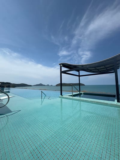 The image displays an outdoor infinity pool at Cenang Beach, Malaysia, under a bright blue sky with scattered cirrus clouds. The pool's surface is tiled with small, light blue square tiles, containing clear water exhibiting minor surface ripples. No individuals are present. A curved, metallic handrail is positioned in the foreground, and a straight metallic handrail is visible mid-ground. To the right, an elevated, smaller rectangular water feature with dark blue water, consistent with a jacuzzi, is partially shaded by a black, four-post structure with a curved, dark roof. A similar partial structure is visible on the far left. The main pool's infinity edge extends towards a tranquil turquoise ocean. Multiple green, tree-covered islands or landmasses are visible on the distant horizon. The scene is illuminated by direct sunlight.