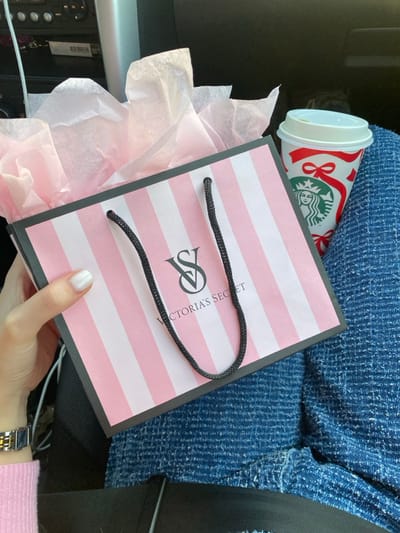 The scene depicts a close-up, downward-angled view inside a vehicle, located in the city of Călărași, Romania. A person's left hand, with white painted fingernails and a silver and gold wristwatch, holds a Victoria's Secret shopping bag. The bag features vertical pink and white stripes, black rope handles, and the "VS Victoria's Secret" logo in black. Light pink tissue paper is visible crumpled at the top opening of the bag. To the right of the bag, a Starbucks paper cup with a white lid rests against a textured blue fabric, consistent with clothing. The cup displays a red and white holiday design with bows and ribbons, along with the green Starbucks siren logo. In the background, elements of a vehicle's interior, including dark panels and white wiring, are partially visible. A black strap, likely a seatbelt, crosses the lower part of the frame.
