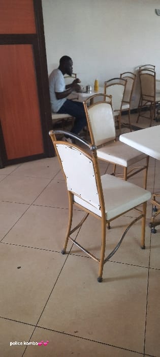 This image depicts an indoor dining establishment, implicitly located in Siaya Township ward, Kenya. One adult male, identified as Black, with dark hair and a beard, is seated at a white-topped table on the left side of the frame. He wears a light-colored short-sleeved shirt and dark trousers, and appears to be eating, holding objects with both hands over a bowl or plate. On his table, a yellow squeeze bottle and a silver cylindrical dispenser are visible.
The room features light-colored, possibly beige or cream, tiled flooring with dark grout lines. The walls are plain and light-colored. To the left, a dark brown wooden door frame or panel is partially visible. The dining area contains multiple empty tables with white tops and chairs with gold-colored metal frames and light-colored, possibly white or cream, upholstered seats and backs. One such chair is prominently featured in the foreground, with its back facing the viewer. The lighting is bright and even, illuminating the entire scene. No other individuals are present in the visible area.