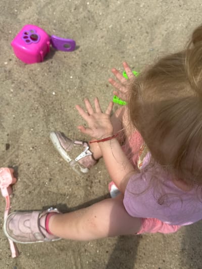 A young child with blonde hair is sitting on a sandy surface. The child is wearing a pink t-shirt and pink shorts. One shoe, a pink sandal, is visible on their left foot, and the other is on the ground. Several toys are scattered around: a pink and purple plastic toy, a green plastic toy, and a pink plastic shovel. The scene appears to be outdoors, possibly in a park or playground, given the sand and toys. There are no other people or identifiable structures in the immediate vicinity. The lighting suggests it is daytime.