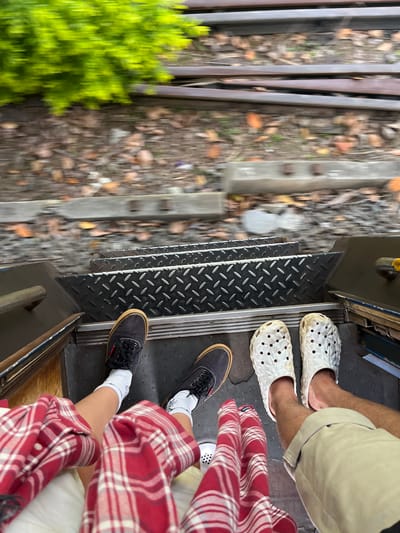 The image shows a top-down view of two pairs of feet standing on a metal staircase. The staircase is diamond-plated and appears to be on some form of transportation, possibly a train or tram, as suggested by the blurred railway tracks and foliage in the background. One person is wearing dark canvas shoes with white socks, and the other is wearing white Croc-like shoes. Both individuals are dressed in casual clothing, with one person wearing a red and white plaid shirt. The scene appears to be outdoors in a motion blur, indicating movement. Based on the provided context that the image is from Kalutara, Sri Lanka, it is likely that this scene is depicting a journey on a local train or possibly a tuk-tuk or other open-air vehicle, with the blurred background suggesting the lush greenery and tracks common in the region.