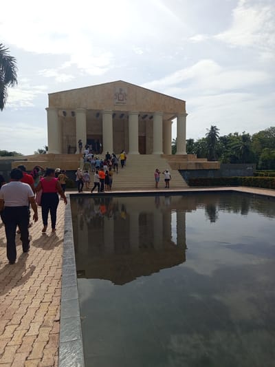 The image shows the entrance to the monument of Miguel Grau in the city of Coveñas, Colombia. A group of people are walking up the stairs to enter the monument, which is a large building with columns and a triangular roof. In the foreground, a paved walkway leads to a large rectangular pool of water that reflects the monument and the sky. The sky is partly cloudy, and there are palm trees in the background.