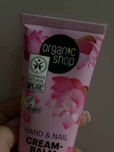 The image shows a hand holding a tube of hand and nail cream. The tube is pink and decorated with images of pink lotus flowers. A black circular logo with "organic shop" written in white is visible on the tube. There is also a rectangular label that says "COSMOS NATURAL 98% NATURAL ORIGIN OF TOTAL" and a circular "Vegan" logo. The text "HAND & NAIL CREAM - BAL" is also visible on the tube. The background is a plain grey surface, suggesting the photo was taken indoors. The lighting is artificial, and the focus is on the product being held. The overall impression is that of a product advertisement or review.