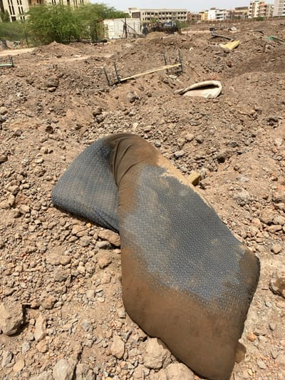 The image shows a large, dirty, dark blue and brown fabric object lying on the ground. The fabric object appears to be a rolled-up mattress or a heavy duty tarp. It is lying on a ground surface composed of dry, rocky soil.  The location is a construction site or a recently cleared area in the city of Khartoum, Sudan, at coordinates 15.528 latitude, 32.571 longitude. The ground is covered in scattered rocks and debris. There are no people or other objects in the scene. The image is taken from an angle above the fabric object. The sky is not visible in the image. The scene appears to be in a dry, dusty environment.