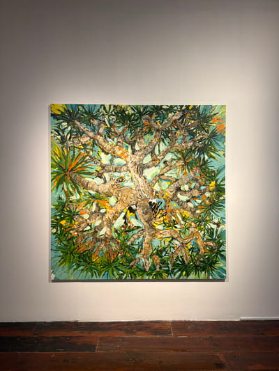A square, textured oil painting of a tree viewed from below is displayed on a white wall, illuminated by a spotlight. The artwork depicts a dense canopy of leaves and thick, twisting branches against a bright turquoise sky. The leaves are rendered in various shades of green, with accents of yellow, orange, and red, suggesting sunlight filtering through. The tree's trunk and branches are painted with thick impasto strokes, creating a tactile surface with a palette of browns, grays, and whites. The composition is dynamic, with the branches creating an intricate, almost abstract pattern.
To the right of the painting, a section of a rustic, dark wooden door is visible, with vertical planks showing a weathered texture and varied wood grain. The wall on the left of the painting is a neutral white, subtly lit to highlight the artwork. The overall impression is of a vibrant, close-up view of nature, likely captured in a warm climate given the style of foliage. The lighting suggests an indoor setting, possibly an art gallery or a well-lit exhibition space. No people are visible in the frame, and there is no visible text. The time of day and weather are implied by the bright, clear sky depicted in the painting. The location context of La Cuesta, Spain, would support the subject matter of a tree with such foliage.