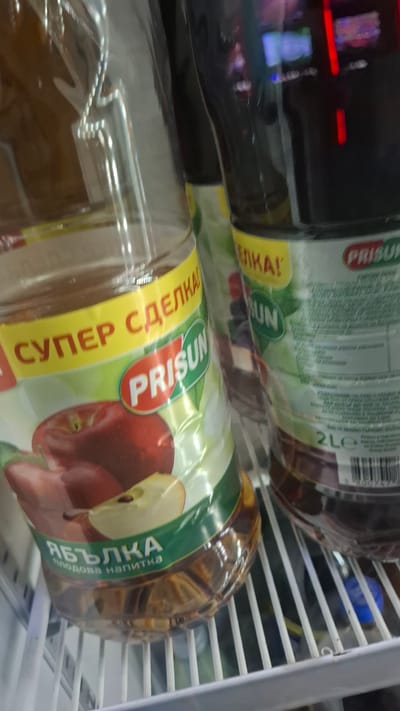 The image shows two plastic bottles of "PRISUN" brand juice drink in a refrigerator. The front bottle is apple flavored and the label has the words "СУПЕР СДЕЛКА!" in yellow. The bottles are stacked on their sides, partially obscuring the bottles behind them. The image was likely taken in Nikopol, Bulgaria, due to the Cyrillic text on the labels.