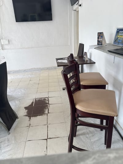 The image depicts an interior scene in Kavrakirovo, Bulgaria. The scene includes a section of a wall-mounted television, three chairs upholstered in a light color with dark wood frames, and a section of tiled flooring with a dark stain. The scene also includes a wall-mounted countertop with a computer monitor and a book on top. The interior space has white walls.