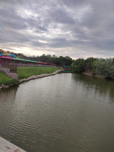 The image captures a scenic view of a park area in Shcherbani, Ukraine. A wide body of water, possibly a river or lake, dominates the foreground, its surface rippling gently under a cloudy sky. The water appears murky green. On the left bank of the water, there is a grassy slope leading up to a paved area with a fence. Behind the fence, there are colorful structures that appear to be part of a water park, with bright yellow, pink, and blue slides visible. Further back, a line of trees forms a dense backdrop. The right bank of the water is also lined with trees, with some lighter foliage interspersed among the darker green. A small, covered structure is visible in the distance on the right bank, nestled among the trees. The sky is overcast with dramatic gray clouds, with patches of lighter sky suggesting diffused sunlight. A few birds are visible in the sky. The overall impression is a tranquil, somewhat overcast day at a recreational park.