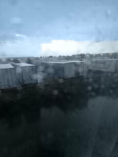 The image shows a view of a logistics company's yard in Uithoorn, Netherlands, on a cloudy and rainy day. Several white refrigerated trailers are parked in rows. One trailer has "BALKENENDE" and "Fresh & Perishable Logistics" printed on its side. Another trailer displays "Yusen Logistics" with a logo. In the background, buildings with lit windows are visible, suggesting a business complex. The foreground is dominated by a dark, wet surface, possibly a road or pavement, with numerous water droplets, indicating rain. The sky is overcast with diffused light.