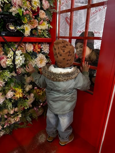 The image displays an artistic installation set within a red structure, reminiscent of a traditional phone booth. The space is filled with a dense arrangement of artificial flowers and green foliage in various shades of pink, cream, yellow, and red.
A child, wearing a light green hooded jacket with a faux fur trim, blue jeans, and brown boots, is positioned horizontally within the booth, reaching their hand into the mass of flowers. They also wear a brown cap with a checkered pattern. An antique-style black rotary telephone with a coiled cord is partially visible among the flowers in the lower-left corner.
Above the child, an adult's face is partially seen through a red grid or grate, looking down into the booth. The adult has a neutral expression and is wearing a dark head covering. Behind this grate, newspaper pages with Russian text are plastered, forming part of the background. Visible Russian words and phrases from the newspapers include "СОВЕТ МИНИСТРОВ ЦЕНТРА" (Council of Ministers of the Center), "Президиуму Верховн" (to the Presidium of the Supreme), "Татарской" (Tatar), and "Автономной Совет" (Autonomous Council). Numeric sequences like "2980" and "2L9S" are also present.
The scene conveys a playful and curious atmosphere, with the child exploring the vibrant floral display. The setting appears to be an indoor or protected environment, possibly in Korobovo, Russia, given the contextual information and visible Russian text.