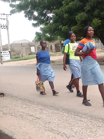 Three young women are walking along a road in Biliri, Nigeria. They are wearing light-colored skirts and carrying items. One is in a blue top and skirt, holding a tote bag. Another wears a neon yellow shirt with a black bag. The third is in an orange and red shirt with a striped skirt. A man is seen in the background, standing near a building with a sign reading "MAI ANGANJE PALACE". A large tree is visible behind the women. The road appears unpaved and dusty. Overhead electrical lines are visible on poles in the background. The buildings are simple concrete structures.