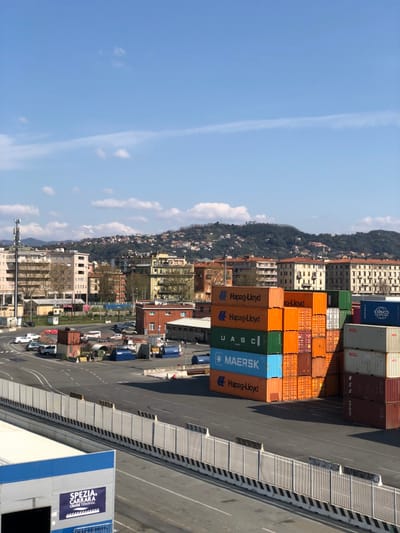 An overhead view captures a port and industrial zone in La Spezia, Italy, under a clear blue sky with sparse white clouds. In the lower left foreground, a segment of a building with a blue and white facade is visible, displaying "SPEZIA CARRARA CRUISE TERMINAL" on a sign. A wide asphalt road with white and yellow lane markings occupies the lower frame, separated from the midground by a grey metal barrier with black and white striped concrete bases.
Beyond this barrier, multiple stacks of shipping containers are present. A primary stack on the mid-right features orange, green, and blue containers, branded "Hapag-Lloyd," "UASC," and "MAERSK," stacked approximately five to six units high. To its right, additional stacks of white, beige, and dark red containers display "COSCO" and "OOCL" logos.
The midground consists of a paved industrial yard containing several parked white cars and vans, arranged along a curved road. Large cylindrical objects, smaller container stacks, and various industrial equipment are distributed throughout. Low-rise industrial buildings with reddish-brown roofs are visible within this area.
The background transitions into a dense urban landscape composed of multi-story residential and commercial buildings, primarily light-colored with terracotta roofs. These structures extend to the base of green hills, which are dotted with scattered houses and exhibit moderate vegetation. No individuals are discernible within the scene, and no active human interactions or tasks are depicted.
