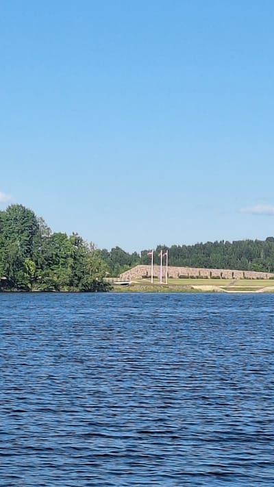 The image captures a serene daytime scene on a body of water, likely a lake or river, in the vicinity of Secē, Latvia. The foreground is dominated by the rippling surface of the deep blue water, reflecting the clear blue sky. In the middle ground, a lush green treeline lines the left bank of the water, with dense foliage creating a natural barrier. Beyond the trees, a stone structure rises, resembling a tiered wall or embankment, possibly a man-made fortification or landscape feature. Three tall flagpoles stand prominently on this structure, each adorned with two flags, one white and one red, suggesting a national or ceremonial significance. The background is a soft blur of more trees and a faint hint of clouds in the sky, completing the tranquil, natural setting.