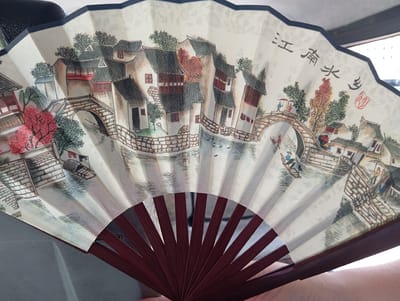 An open folding fan displays a painted scene reminiscent of a traditional Chinese water town. Buildings with dark tiled roofs and light-colored walls line canals crossed by arched stone bridges. Figures are depicted in the scene, including people on the bridges and in boats on the water. Chinese characters and a red seal are visible in the upper right corner of the fan. The fan's ribs are made of dark wood. The scene is not located in Yukariyuz, Uzbekistan, but rather depicts a landscape found in regions of China known for water towns, such as Jiangnan.