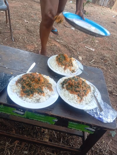 An outdoor scene in Egede, Nigeria, depicts three white round plates of food served on a dark brown wooden table or bench. Each plate contains a mound of white rice topped with a stew or sauce featuring reddish-orange broth, green leafy vegetables, and small protein pieces. Metal spoons rest on or beside two of the plates. A person with dark skin tone, wearing blue flip-flops on one foot, stands behind the table, holding an oval blue and silver platter with their right hand. The ground is visible behind the table, consisting of dry dirt and sparse foliage. A metal chair leg is partially visible to the left. Green promotional stickers are affixed to the underside wooden frame of the table. A crumpled silver foil wrapper lies on the table to the right.