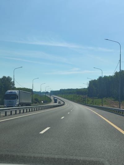 A daytime, eye-level shot shows a highway in Shchedrovka, Russia, on a clear day. The sky is bright blue with wispy white clouds. The road curves to the right, with guardrails lining both sides. Trees and green hills form the background. Several vehicles are visible: a white semi-truck with a trailer is in the left lane, and further ahead, two other trucks and a car are in the distance on the right lane. Streetlights line the highway. The road surface is asphalt, with white lane markings and a yellow line dividing the lanes. The scene appears to be a typical highway commute in a rural or suburban area.