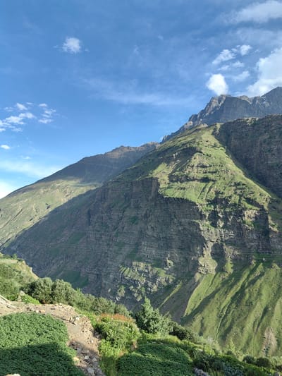 The image depicts a vast mountainous landscape in Tiling, India, under a clear blue sky dotted with white clouds. The foreground features lush green vegetation and rocky terrain. Dominating the scene are imposing, steep mountainsides covered in vibrant green grass and characterized by rugged, layered rock formations. The sunlight casts dramatic shadows across the slopes, highlighting the topography. Power lines are visible stretching across the mountains, indicating human presence and infrastructure within this natural environment. The overall impression is one of rugged natural beauty and grandeur.