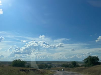 The image depicts a rural landscape in the village of Lopățica, Moldova. A paved road curves through dry, grassy hills dotted with trees. Two vehicles are visible on the road: a dark SUV and a white crossover. The sky is a vibrant blue, filled with fluffy white cumulus clouds. In the distance, rolling hills suggest agricultural fields. The overall impression is one of a sunny day in the Moldovan countryside.