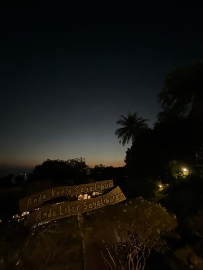 The image depicts a dusk or twilight scene in Karimunjawa, Indonesia. Two wooden signs are prominently displayed in the foreground. The top sign reads "Anda Kerja Saya Libur," and the bottom sign reads "Anda Libur Saya Liburan." These phrases translate to "You Work I Vacation" and "You Vacation I Vacation." Behind the signs, silhouetted trees and foliage create a natural backdrop, with a palm tree visible to the right. In the distance, faint lights suggest a settlement or buildings. The sky transitions from a deep blue to a lighter hue near the horizon, with a hint of orange from the setting sun. The overall atmosphere is serene and suggests a holiday or leisure destination.