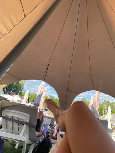 In Zazymia, Ukraine, this outdoor scene depicts individuals relaxing under a large, taut fabric canopy. The canopy, a neutral beige color with dark trim, is supported by a central pole and features a segmented design. Through an arched opening in the canopy, a bright blue sky with scattered white clouds is visible, along with a line of lush green trees and buildings. In the foreground, a person's legs and feet are prominent, suggesting they are reclining on a lounge chair. In the background, several closed beach umbrellas stand in a row, and other lounge chairs are visible. A person's legs are visible on a chair in the middle ground, and a can is on a table nearby. The overall impression is one of leisure and outdoor relaxation.