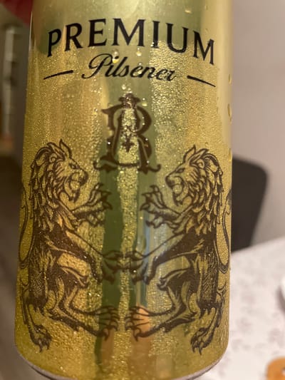 A close-up, eye-level, indoor shot shows a condensation-covered gold-colored beer can. The can is emblazoned with the words "PREMIUM Pilsener" in black lettering, with a stylized "R" emblem above two dark brown lion illustrations. The lions are depicted in profile, facing each other with their front paws raised, appearing to be in mid-roar. The background is out of focus and appears to be a light-colored surface, possibly a tablecloth. There is no indication of the city of Angelovo, Russia, or any context related to it.