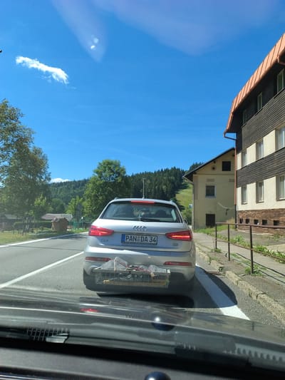 The image depicts a silver Audi Q3 car from the rear, stopped on a road. The car's license plate reads "PAN DA 34". Attached to the rear bumper is a metal basket containing bundled items secured with rope, possibly luggage or other belongings. The car is on a road with a white line marking. To the right of the road, there is a sidewalk and a metal fence, beyond which are buildings with a dark-brown roof and cream-colored walls, and more buildings with dark wooden siding. In the background, there are trees and a forested hill. The sky is clear and blue with a single small cloud. The scene is consistent with a rural or semi-rural setting in Horní Vltavice, Czechia, given the license plate format and typical building architecture. The perspective suggests the photo is taken from another vehicle following the Audi.