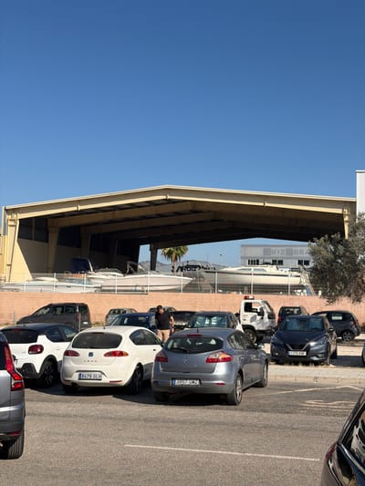 This media file captures a scene in a parking lot outside a commercial building, likely an industrial or business complex. The sky is a clear, bright blue, suggesting a sunny day with good weather. The image is oriented vertically, with the ground and vehicles taking up the bottom two-thirds of the frame and the sky the top third.
The primary objects are several parked cars of various colors, including white, grey, and dark blue. Some of the cars appear to be in a service or repair area, with some of them possibly being worked on or displayed. In the background, a long, low-slung building with a light-colored roof supported by exposed beams is visible. The building has large open areas under the roof, suggesting a workshop, storage, or covered parking. Some windows and signage are visible on the building's facade.
One car, a white hatchback, has a license plate that reads "8476 GLH". Another car, a dark grey sedan, has a license plate "1170 KNF". A person wearing a black t-shirt and beige shorts is visible near the cars, appearing to be observing or interacting with them.
The setting appears to be a sunny outdoor environment, possibly in the afternoon given the quality of light. The presence of multiple vehicles and the industrial-style building suggest a commercial or industrial area. Some palm trees are visible in the background, hinting at a warm climate. The overall impression is one of everyday activity in a business park. There is some text visible on the building which appears to read "RUIZ", and "RUIZ Y GASA" on a fence.