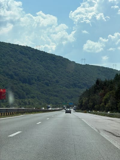This image depicts a highway with a dense, green, forested mountain slope on the left and a clear blue sky with fluffy white clouds above. Several electricity pylons are visible on the mountainside. The highway is paved and has white lane markings. There is a metal guardrail separating the highway from the mountain. On the highway, several vehicles are seen traveling in the same direction, indicating a steady flow of traffic. Among the vehicles, a white car is prominently visible. Blue road signs are present, with some text visible, though it is difficult to read clearly in the overall image. One sign appears to indicate directions with arrows. The scene suggests a daytime setting with fair weather. The presence of a mountain and highway suggests a location that might be mountainous or hilly. The sign indicating "Potop, Bulgaria" provides specific location context. The overall impression is of a scenic drive through a natural landscape with modern infrastructure.