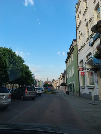 The image captures a view down a cobblestone street in Strakonice, Czechia, lined with buildings and trees. In the foreground, the rear of a silver car is visible. Further down the street, several other cars are parked. The buildings on the right side of the street are multi-story structures with windows, some featuring awnings and signs. A prominent green building is visible, with a Coca-Cola sign hanging on its facade. On the left side of the street, large, leafy trees create a canopy. The sky above is a clear blue with a few scattered white clouds. The scene appears to be set in the late afternoon or early evening, given the lighting conditions.