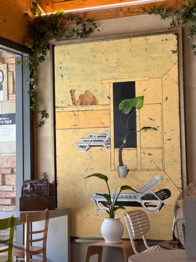 The image depicts an indoor establishment, likely a cafe or gallery, characterized by textured light beige walls and exposed wooden ceiling beams. Artificial green ivy vines are draped decoratively along the wooden beams and down the left wall.
The central and dominant feature is a large, vertically oriented painting within a thin, light-colored frame. The painting features a textured, pale yellow-beige background with a scratchy, distressed appearance, suggestive of an arid environment. Depicted within the painting are: a light brown camel, partially visible from the chest up, positioned towards the upper left, facing right; a stack of three white modern lounge chairs on the mid-left; a single white modern lounge chair on the lower right, casting a dark shadow; a dark, rectangular void resembling a doorway or window, through which a large, green-leafed plant in a grey pot appears to float.
To the left of the painting, a dark vertical beam or frame element is present. Below it, a wooden cabinet or shelf holds several stacked dark rectangular objects, possibly books or CDs, topped by a small, shiny golden bird figurine. Further left, a portion of a window or doorway reveals an exterior brick wall. A white sign with Korean text, including "GIGAeyes TV 작품중" (GIGAeyes TV, currently featuring/showing work), is affixed to the left wall. Partially visible chairs include a green wooden chair and a standard wooden chair in the lower-left foreground.
In the lower-right foreground, a small, light-colored stool or low table supports a white, speckled globular pot containing a green plant with long, dark leaves, resembling a Peace Lily. A light-colored, slatted modern chair is partially visible behind this plant. On the far right, a metallic, standing fan is partially visible. Overhead lighting fixtures are visible along the wooden beams. The scene is static, with no visible individuals. The exact address in Jongno-gu is not discernible from the image, but the presence of Korean text confirms its location in South Korea.