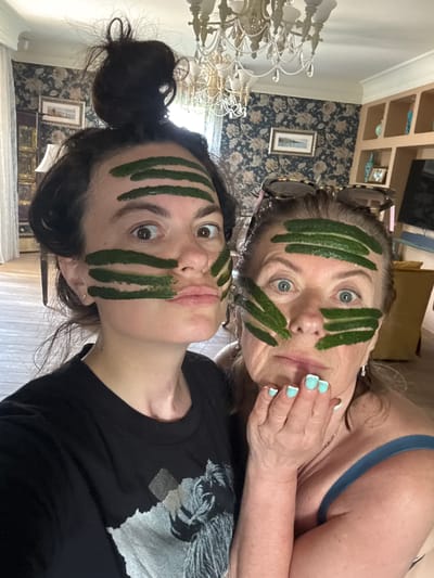 Two women are posing for a selfie indoors, with green face masks applied in horizontal stripes to their foreheads, cheeks, and chins. The woman on the left has her hair in a messy bun and is wearing a black t-shirt. The woman on the right has sunglasses on top of her head and is wearing a blue tank top; her fingernails are painted with a light blue French manicure. The background suggests an indoor setting with floral wallpaper, a chandelier, and built-in shelving.
