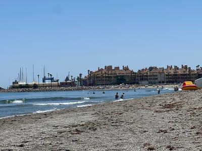 The image shows a beach scene in San Roque, Spain, with the ocean in the foreground and buildings and a marina in the background. The beach is covered in pebbles and seaweed. Several people are in the water, swimming and wading. Some people are on the beach, sunbathing and relaxing. An inflatable water toy, red and yellow, is partially visible on the right side of the image. The buildings in the background appear to be apartments or hotels, with terracotta roofs and palm trees in front of them. The marina features several sailboats and other vessels. The sky is clear and blue, indicating a sunny day.