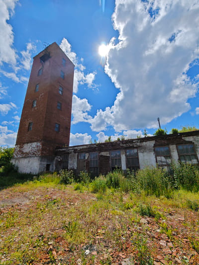 This is a wide-angle, eye-level shot of a dilapidated brick tower and an adjacent low-rise structure, likely an abandoned industrial building, under a bright, partly cloudy blue sky. The sun is visible and glinting, creating a lens flare in the upper right quadrant of the image. The tower, constructed of weathered red brick with white plaster on its lower section, has multiple small, dark-paned windows. The roofline of the low-rise building is overgrown with vegetation, and its windows are broken or boarded up. The foreground is a patch of dry, uneven ground littered with red brick fragments and sparse, dry grass. The scene appears to be located in an open, possibly rural or industrial area outside of a city, with no people or vehicles present.