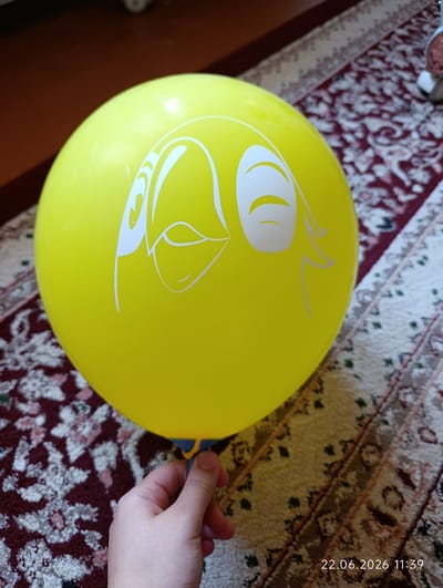 A yellow balloon with a cartoonish white face design is held by a person's hand against a rug. The balloon faces the viewer, featuring large white eyes with curved lids and beak-like markings. The hand, gently holding the balloon's blue and yellow plastic stem, is positioned at the bottom right of the frame. The background is a patterned rug with red and cream colors, and a wooden surface is visible in the upper left corner. The timestamp "22.06.2026 11:39" is present in the lower right corner. The scene is likely indoors, possibly in a residential setting in Eshanguzar, Uzbekistan.