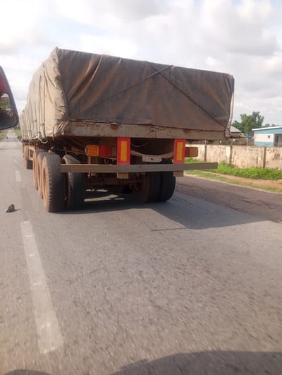 The image shows the rear of a large, covered truck driving on a paved road in Wuro Sani, Nigeria. The truck's cargo is obscured by a brown tarp, which appears worn and dusty. The truck has multiple axles, with several large tires visible. Red and amber lights are mounted on the rear of the truck, likely for signaling. The road has white lane markings.
In the background, there is a low, light-colored concrete wall with green vegetation growing behind it. A building with a blue and white facade can be seen further in the distance, along with trees. The sky is partly cloudy, suggesting it is daytime. The lighting and shadows indicate the sun is out, but the clouds may be moderating the intensity.
The perspective suggests the photo was taken from another vehicle traveling behind the truck, possibly from the passenger seat or side mirror. A small, dark object, possibly a stone, is on the road to the left of the truck. No people are clearly visible, but the context implies travel and transportation. The overall scene depicts a typical roadside view in a rural or semi-urban area of Nigeria.