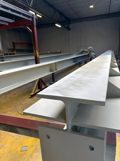 The image depicts an industrial setting, likely a workshop or factory, in Mol, Belgium. In the foreground, a large, light-colored steel I-beam is prominently displayed, supported by a red and yellow metal structure. The beam shows signs of recent painting, with a light, speckled texture. In the background, another similar I-beam is visible, with a person wearing protective gear, including a mask and hood, spray-painting it. The floor is yellow, and the overall environment suggests a manufacturing or construction-related process. The lighting is industrial, with fluorescent lights visible on the ceiling.