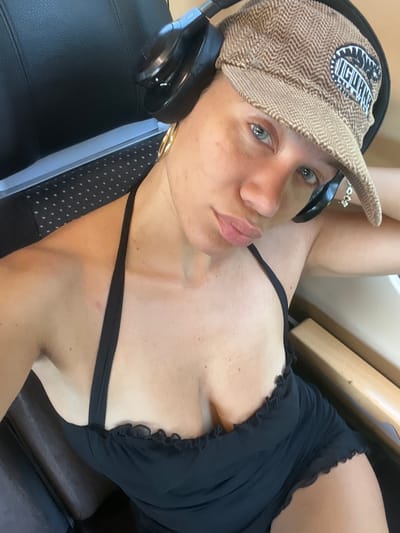 This is a close-up, high-angle, indoor shot of a woman lying down, wearing a tan herringbone patterned baseball cap with a black and white logo patch and a black ruffled bikini top. She is also wearing black headphones. The woman has light skin, blue eyes, and pink lips. A portion of her chest and arm are visible. She is lying on a black surface that appears to be a chair or seat. A light wooden structure is visible in the background. The lighting suggests it is daytime, and the setting is likely indoors, possibly a gym or a lounge area. The woman's expression is neutral. The logo on the hat is difficult to read but appears to contain an animal or mythical creature and some text.