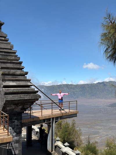 This image, taken in Cemorolawang, Indonesia, depicts a person standing on a viewing platform overlooking a vast, arid landscape. The platform, constructed with a wooden deck and metal railings, juts out from a stone structure with tiered steps, possibly part of a temple or monument. The person, wearing a pink long-sleeved shirt and blue shorts, stands with arms outstretched, holding a water bottle in their right hand. The background features rolling green mountains partially shrouded in mist, under a clear blue sky dotted with white clouds. Trees with feathery foliage frame the right side of the image. In the foreground, shadows suggest the presence of other structures and vegetation.