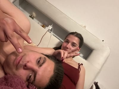 Two individuals are captured in a close-up selfie. The male figure, partially covered by a pink textile, is positioned towards the right side of the frame, while the female is to the left, both striking a peace sign pose. The background appears to be an interior space, possibly a room. There is no readily identifiable evidence to confirm its location in Lüneburg, Germany.