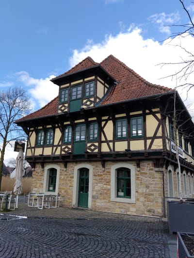 The image displays a multi-story, half-timbered building with a stone foundation, located in Burgsteinfurt, Germany. The structure features a steep, red-tiled gabled roof. The ground floor is constructed from irregular light brown and yellow stone blocks, presenting three arched openings: two windows with green frames and a central green wooden door. The number "17" is visible above the right ground-floor window. A small potted plant is observed within the left ground-floor window.
The upper floors are characterized by a half-timbered construction, featuring pale yellow infill panels and dark brown timber beams with decorative cross-bracing. Multiple rectangular windows with green frames are present on these floors, along with two distinct dark green panels that resemble shutters or doors on the first floor facade. A dark, ornate hanging sign, partially reading "Burg...Stuben", is suspended from a bracket on the building's left side. Another sign displaying "Gastronomie" is visible on the building's right upper section.
In the foreground, a dark grey cobblestone plaza is visible, paved in a radial or fan-like pattern, with noticeable wet patches. On the left side of the plaza, several light-colored outdoor tables and chairs are arranged near the building, alongside a large, folded, light-colored patio umbrella. Tall, dry grasses or reeds are visible behind the outdoor seating. Bare deciduous trees are present on both the left and right sides of the scene, indicating a dormant season. The sky is partly cloudy, with scattered white clouds against a blue background. No individuals are present, and no actions or interactions are occurring.