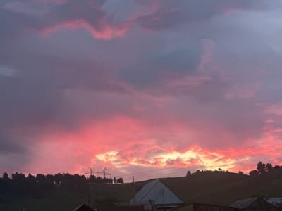 A sunset is depicted in the sky over Aumyshevo, Russia, casting vibrant pink and orange hues across the clouds. Below the colorful sky, a silhouette of trees lines a rolling green hill. A large, lattice-like power line tower stands prominently on the hill, with wires extending across the scene. In the foreground, the roofs of several buildings are visible, suggesting a rural or semi-rural setting. The overall atmosphere is serene, with the dramatic sky dominating the landscape.