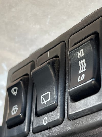 This is a close-up, eye-level, indoor photograph of a section of a black plastic control panel. The panel contains three buttons, arranged in a row from left to right. The leftmost button has an icon of a windshield wiper with a single line above it. Below that is another icon depicting a windshield wiper with a spray of water. The middle button is larger and rectangular, with an icon of a windshield wiper viewed from the side. Below this button is a circular depression labeled with the letter "O". The rightmost button is also rectangular and has white text indicating "HI" at the top and "LO" at the bottom, separated by a symbol of three wavy lines, suggesting heat settings. The background appears to be a light-colored, textured surface, possibly a wall or ceiling. There are no people or animals visible in the image, and no evidence to suggest the location is within the city of Lanžhot, Czechia. The image focuses solely on the control panel and its buttons.