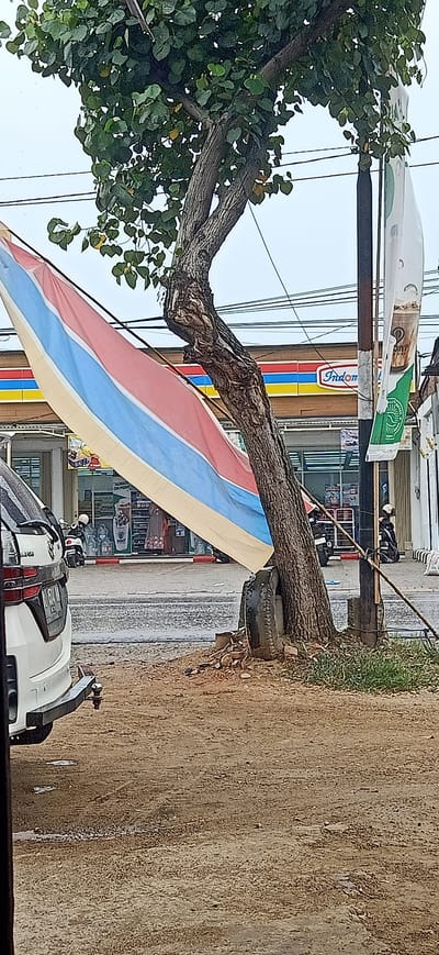 The image depicts an outdoor scene in Uteungeulinggang, Indonesia, on a cloudy day. A large tree with a gnarled trunk stands prominently in the foreground, with its branches extending upwards and its leaves creating a dense canopy. A wide, multi-colored banner, featuring horizontal stripes of blue, red, and yellow, is draped over a branch and extends diagonally across the image. A portion of a white vehicle is visible on the left side, with its rear bumper and hitch in frame. Behind the tree, a building with a distinctive red, blue, and yellow striped facade, identified by the "Indomaret" logo, suggests it is a convenience store. Several motorcycles are parked in front of the store, and some individuals in helmets can be seen. A dark pole, likely a utility pole, is situated to the right of the tree, with wires running overhead. A portion of another white banner with green accents is attached to the pole. The ground in the foreground is a mixture of dirt and gravel, with some scattered debris and patches of green grass. The overall impression is a street view of a commercial area with a natural element in the form of the tree.