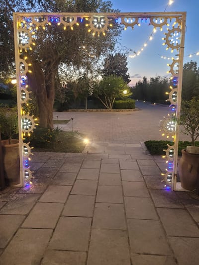 This image, taken in Sogliano Cavour, Italy, captures a charming outdoor scene at twilight or early evening. The primary subject is a decorative, white rectangular archway, richly adorned with numerous circular, spoked light fixtures. These fixtures, resembling ship's wheels or stylized suns, hold a mix of glowing white and warm yellow bulbs, interspersed with bright blue lights. Additional string lights with smaller bulbs are draped across and through the archway, further enhancing the festive atmosphere.

The archway frames a wide, paved pathway made of large, light-colored rectangular pavers, extending into the background. On both sides of the path, lush green trees and bushes are visible, with a prominent, large-trunked tree (possibly an olive tree) on the left. Subtle ground-level lights illuminate sections of the path and surrounding foliage. Rustic, large terracotta pots containing plants are visible on the right side near the archway. The sky above is a clear, deep blue, indicating the transition from dusk to night. The scene suggests an inviting entrance or a decorative pathway within a garden, outdoor venue, or private estate, designed to create a warm and enchanting ambiance. No people or visible text are present in the image.
