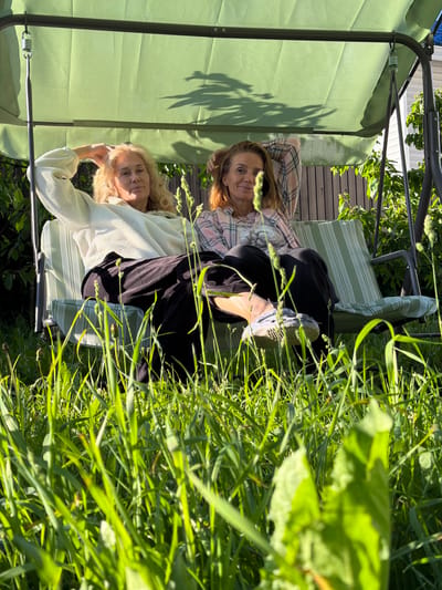 Two women are seated on a green-striped swing set in a garden. The woman on the left has blonde hair and is wearing a light-colored fuzzy sweater and black pants. She has her left arm resting on the back of the swing and her right hand on her head. The woman on the right has reddish-brown hair and is wearing a pink and white plaid shirt. She has both arms behind her head. The swing set is positioned under a green canopy and is surrounded by lush green grass and foliage. The background shows a wooden fence and some white siding, suggesting a residential area. The lighting indicates it is daytime with bright sunlight casting shadows. The overall scene depicts a relaxed moment outdoors.
