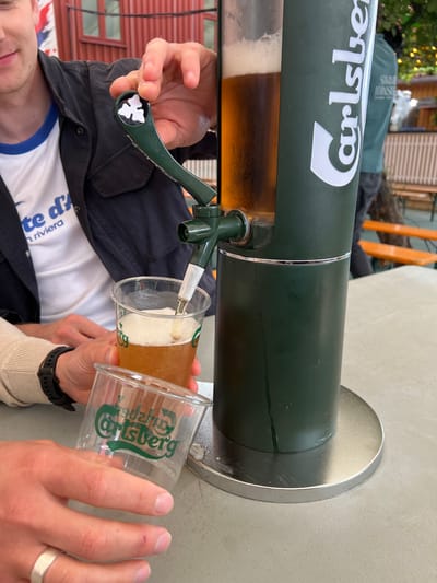 This is a close-up, eye-level shot of a person's hands operating a green beer dispenser with the "Carlsberg" logo prominently displayed. The dispenser is dispensing amber-colored beer into a clear plastic cup, creating a foamy head. Another empty plastic cup with the "Carlsberg" logo is held by a person's hand in the foreground. The dispenser is located on a table, possibly outdoors, with a blurred background that suggests a public area with wooden benches and a building with red siding. The overall impression is of a casual social setting where drinks are being served. The scene implies that this is likely occurring at an establishment in Tønsberg, Norway, given the presence of the Carlsberg branding, a popular beer brand often associated with European establishments.
